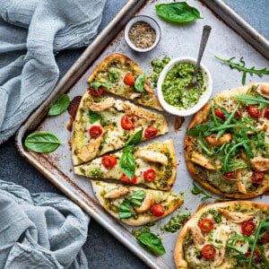 Naan pizza triangles on a baking sheet with fresh basil leaves and arugula around, and pesto in a small white bowl.