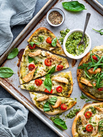 Naan pizza triangles on a baking sheet with fresh basil leaves and arugula around, and pesto in a small white bowl.