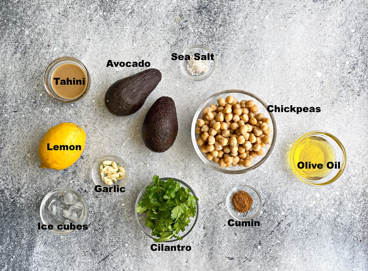 Recipe Ingredients: whole avocados, lemon juice, tahini, salt, cumin, oil, garlic, and chickpeas on a gray background. 