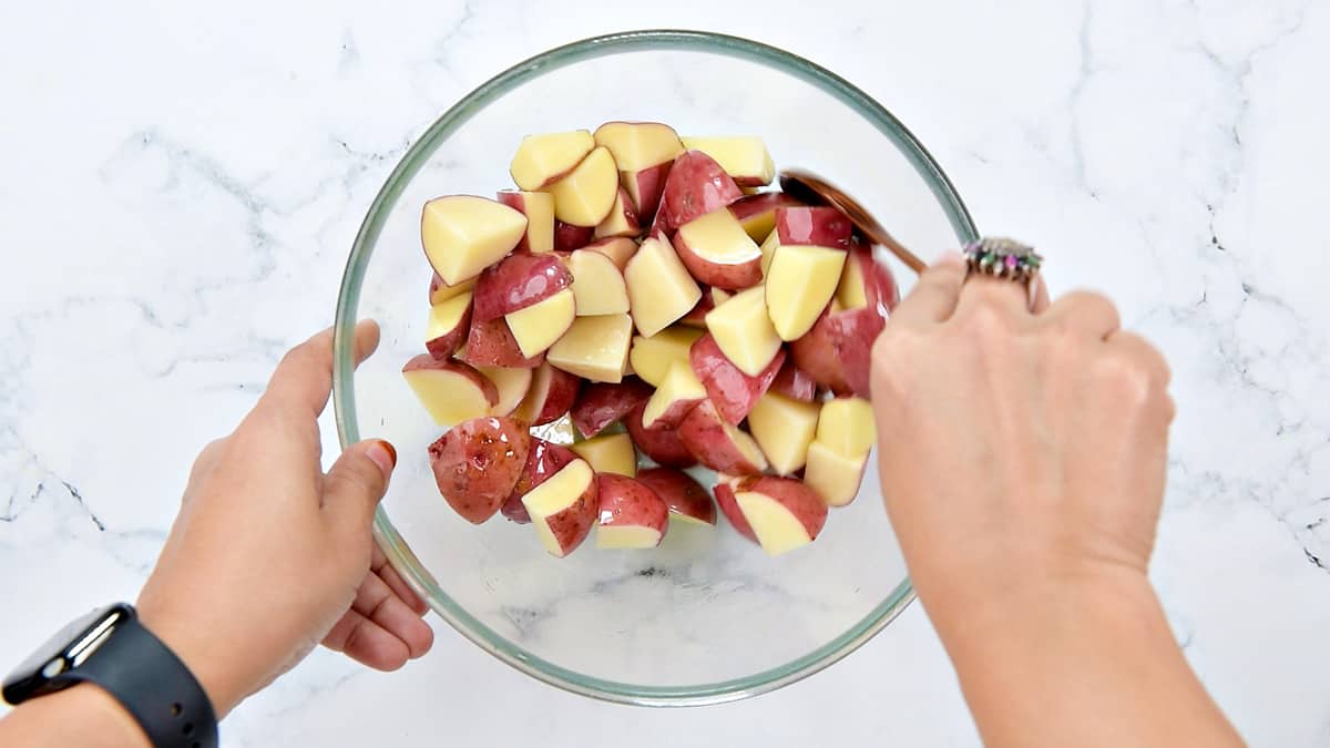 Hands tossing red potato cubes in a glass bowl.