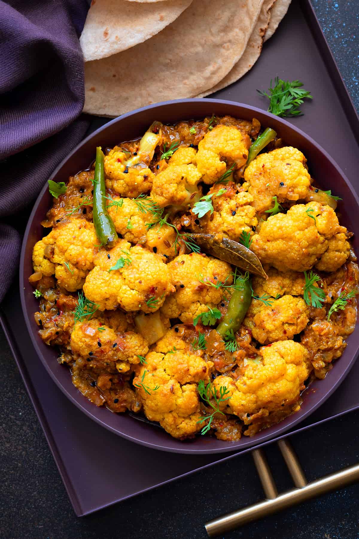 close-up view of achari gobi sabzi served in purple plate with roti on side.