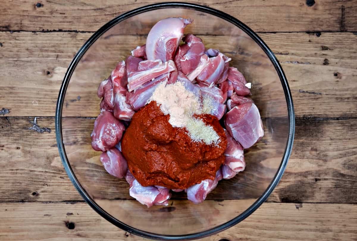 Bone-in lamb pieces, vindaloo paste, sugar and salt placed in a glass mixing bowl.