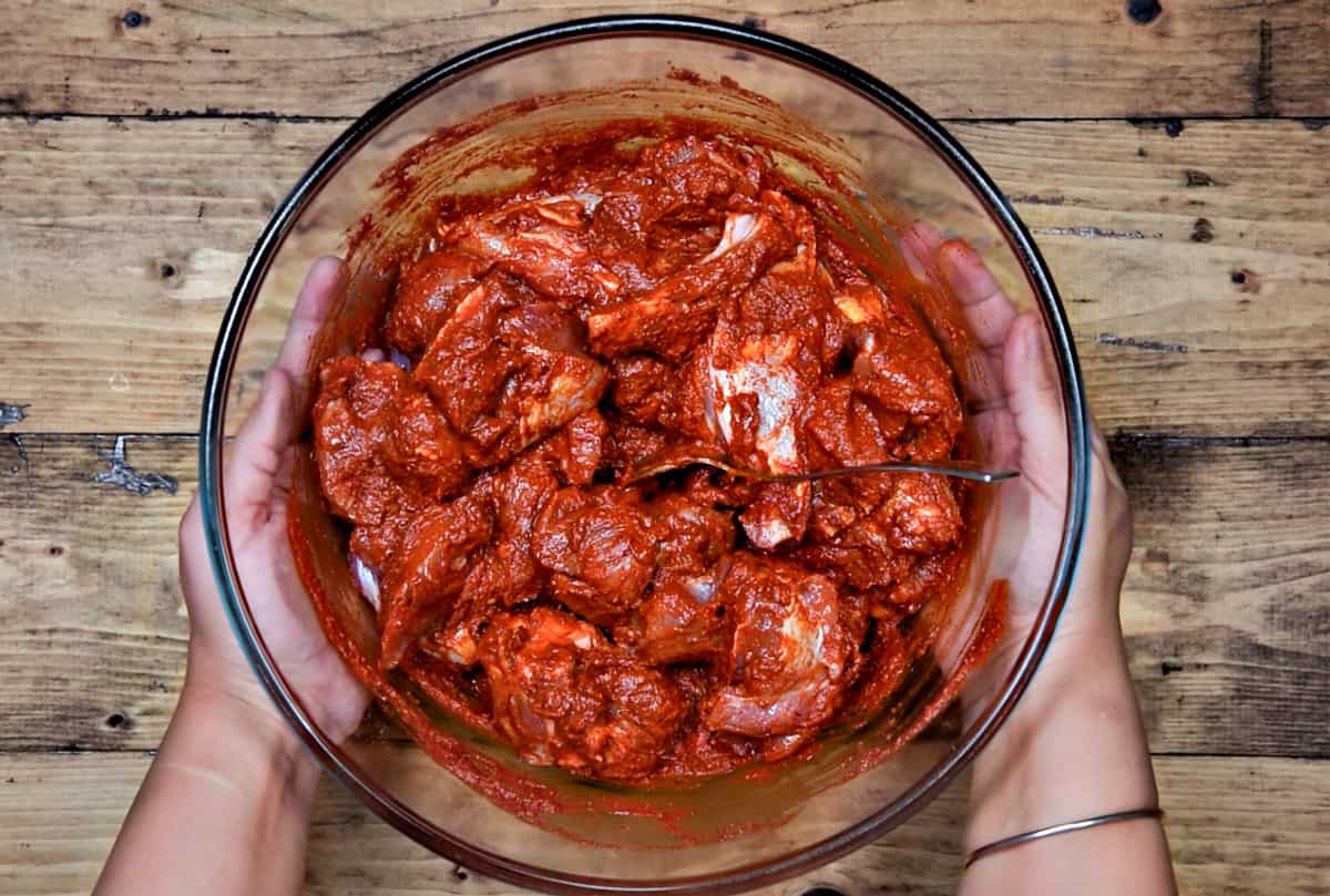 Two hands holding the glass bowl with lamb meat mixed with vindaloo paste. 