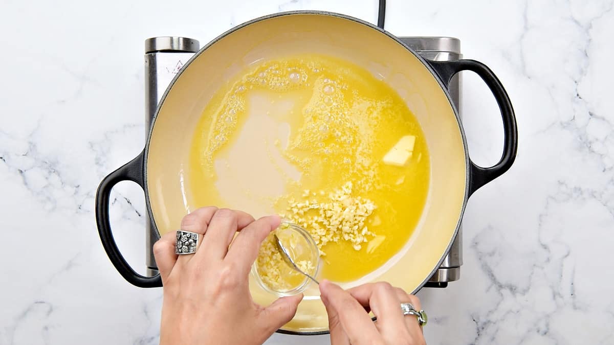 Hands adding minced garlic to melted butter in a cooking pot.