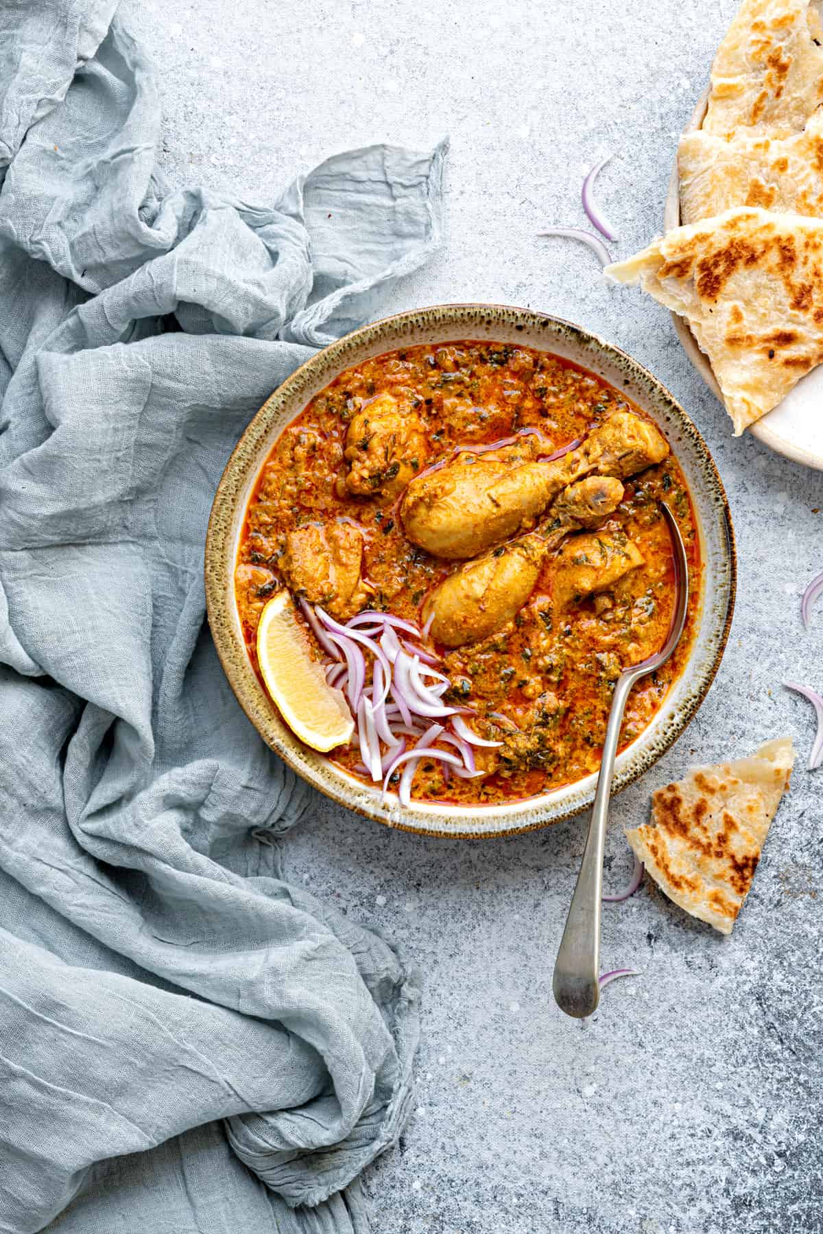 Fenugreek Chicken curry served in ceramic bowl with a serving spoon into it and parathas on side.