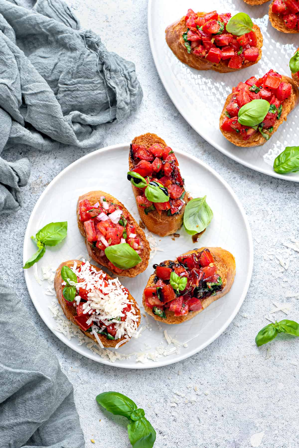Top down view of a white plate with 4 servings of bruschetta. A few pieces of baby basil garnish the plate. One of the baguettes with tomatoes is topped with shredded parmesan cheese.