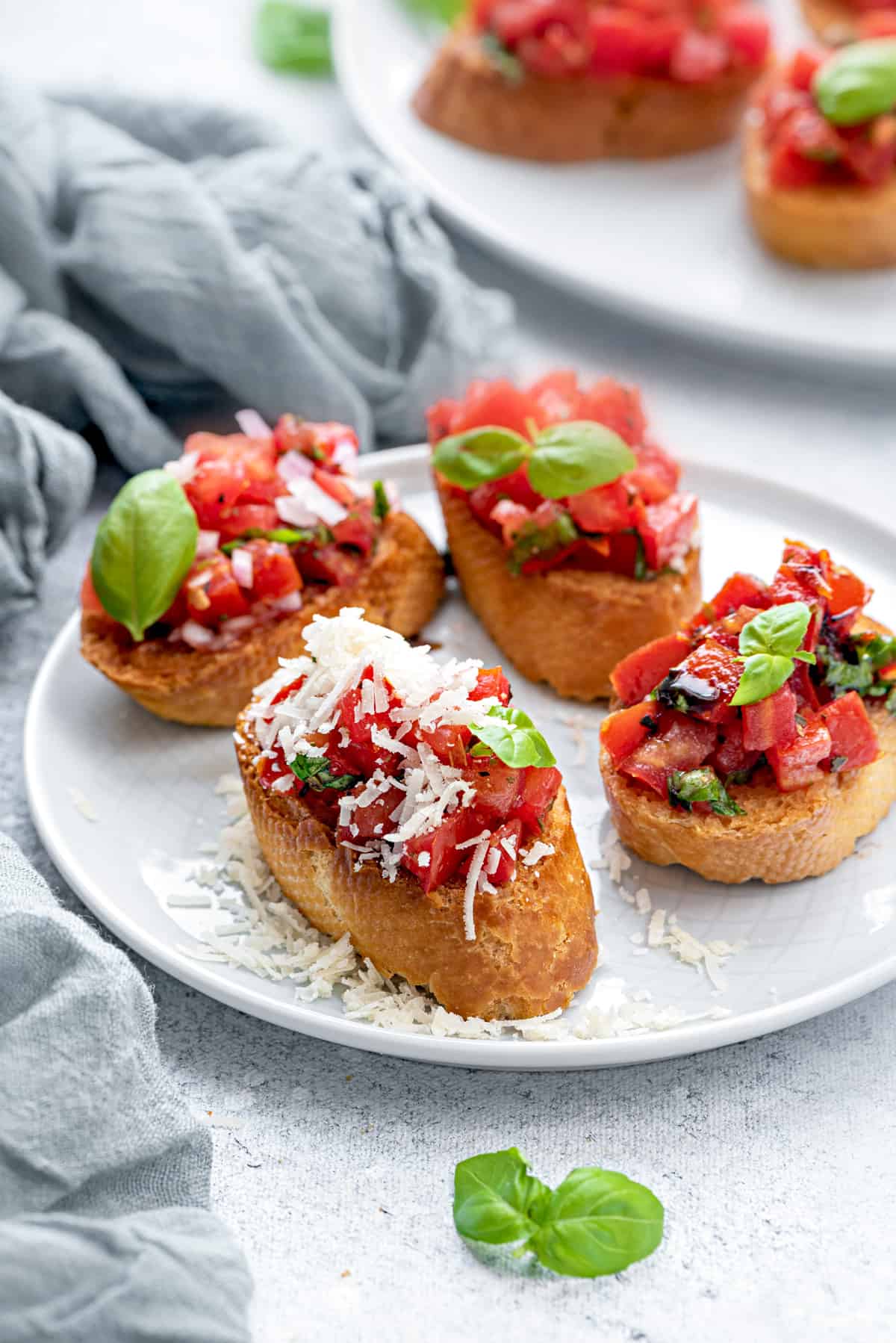 4 servings of bruschetta. A few pieces of baby basil garnish the plate. One of the baguettes with tomatoes is topped with shredded parmesan cheese.