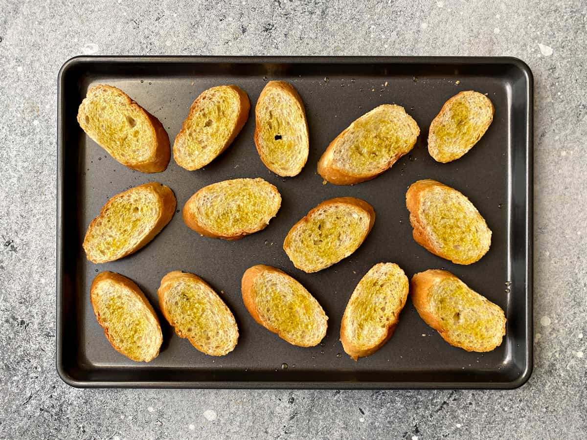 14 pieces of baguette slices drizzled with olive oil and laid on a baking pan.