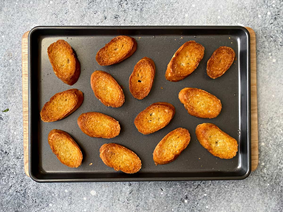 14 pieces of toasted baguette slices on a baking pan.
