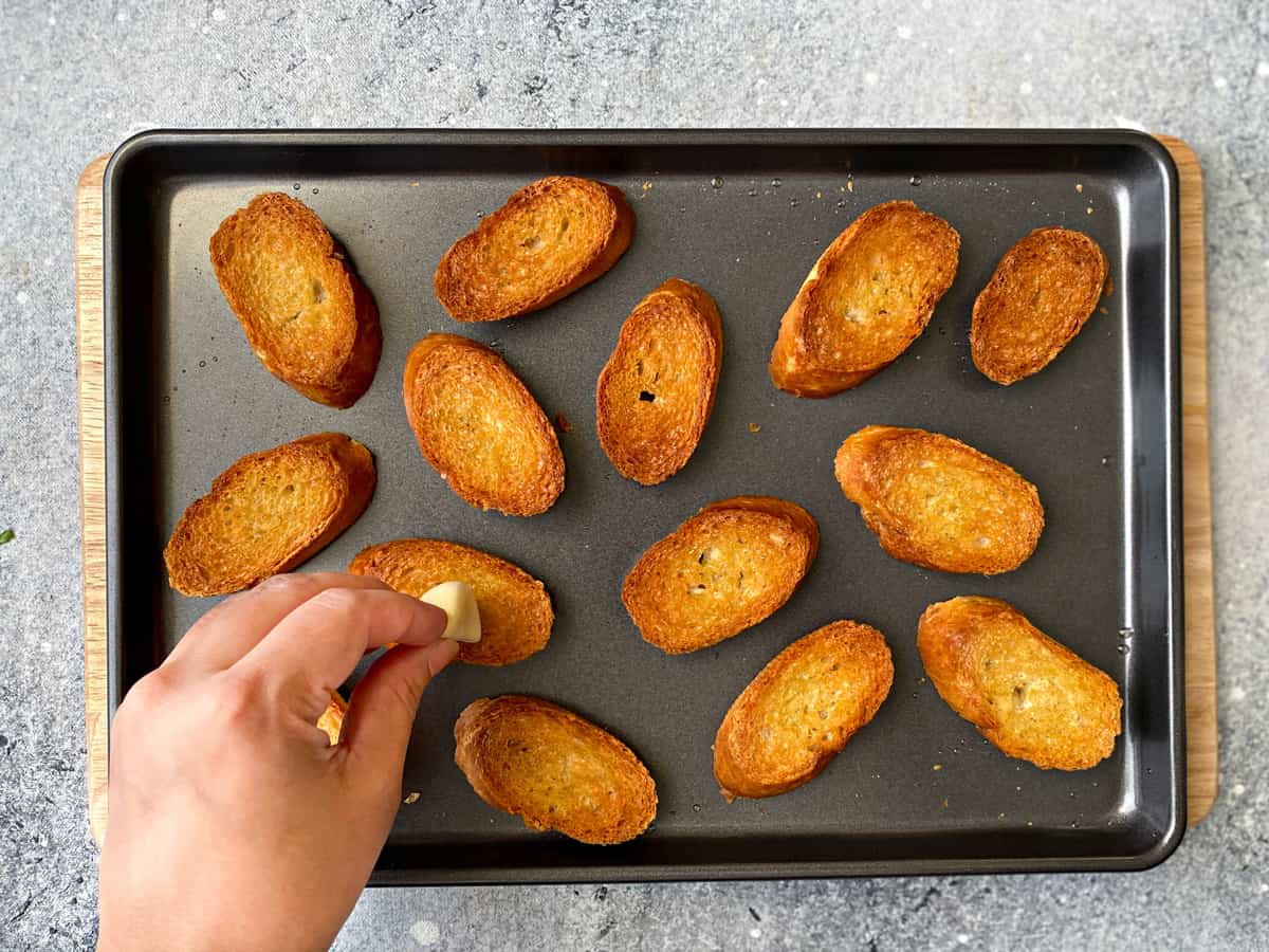 14 pieces of toasted baguette slices on a baking pan. A hand is rubbing half a clove of garlic across one piece of toast.