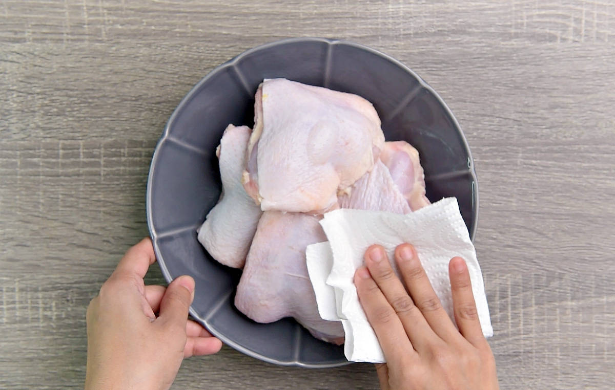 Hand using paper towel to blot excess moisture from bowl of raw skin-on chicken thighs.
