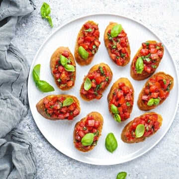Top down view of a white plate with 10 servings of bruschetta. A few pieces of baby basil garnish the plate.