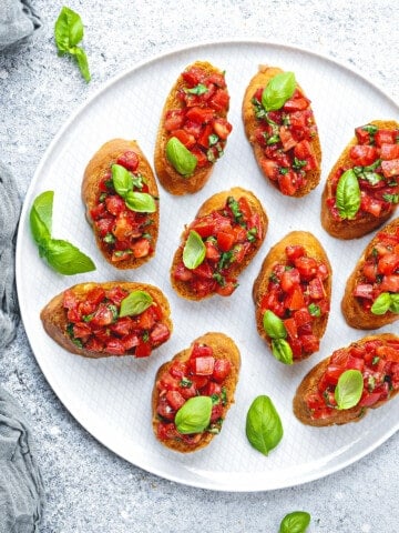 Top down view of a white plate with 10 servings of bruschetta. A few pieces of baby basil garnish the plate.