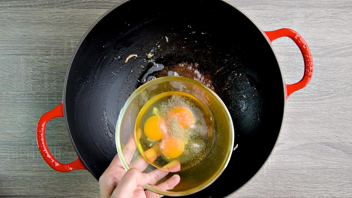 Hand pouring 3 eggs from a glass bowl into a wok.