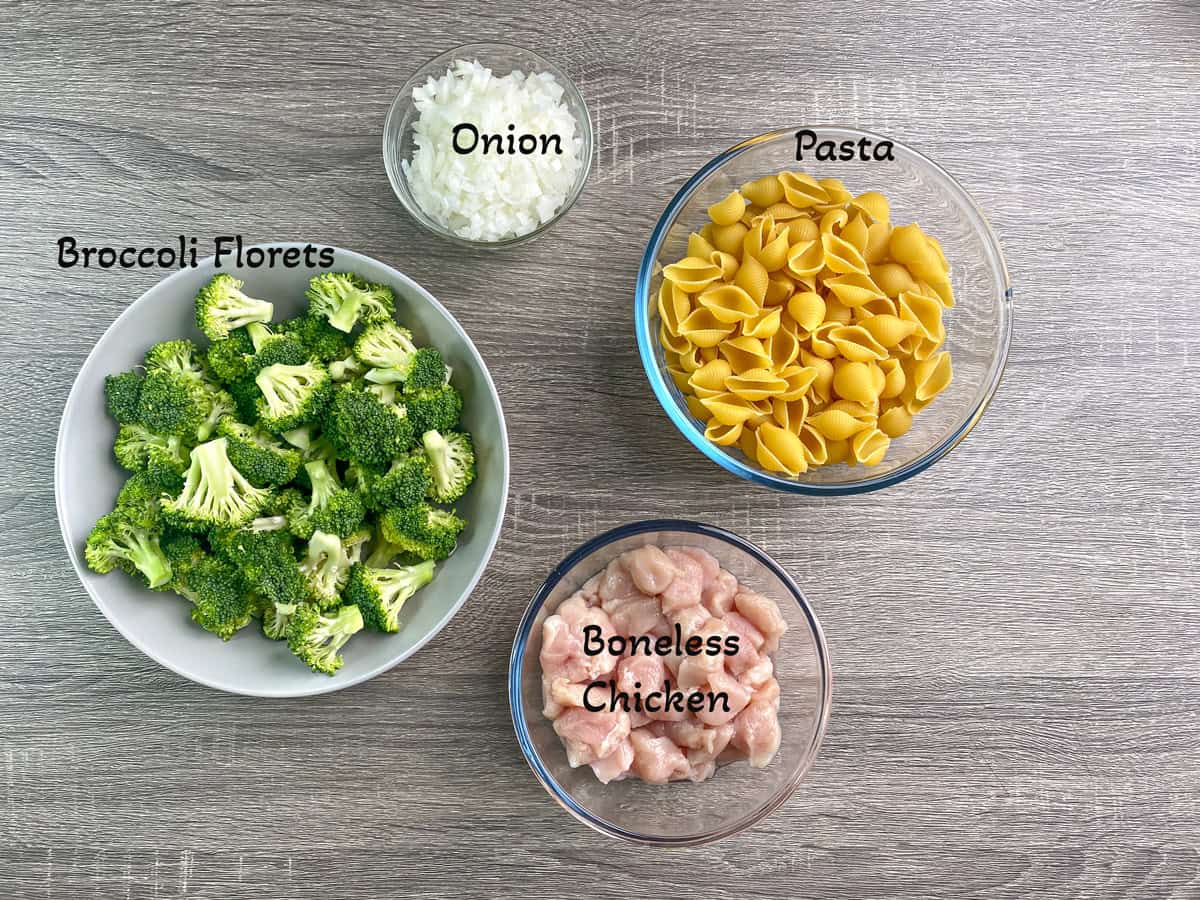 Recipe ingredients: ingredients in individual glass containers. Pasta, chopped onion, broccoli florets, and cubed chicken breasts.