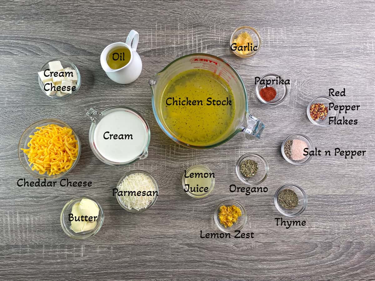 Recipe ingredients in individual glass dishes. Chicken stock, cream cheese, shredded cheese, cream, butter, parmesan, lemon juice and zest, oregano, thyme, salt and pepper, paprika, chili flakes, and oil.