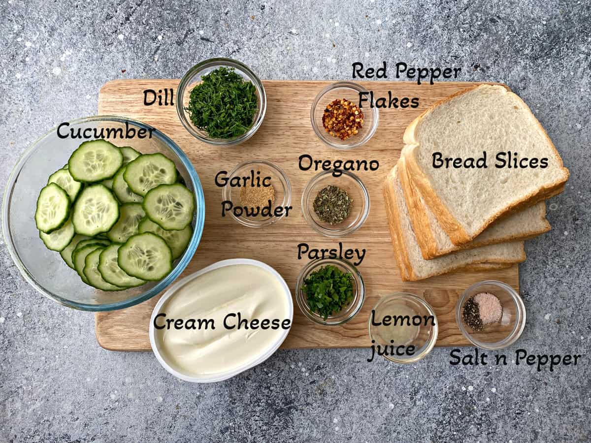 Recipe ingredients on a cutting board - White bread slices, cream cheese container, and sliced cucumbers, fresh dill, parsley, seasonings in individual glass bowls.