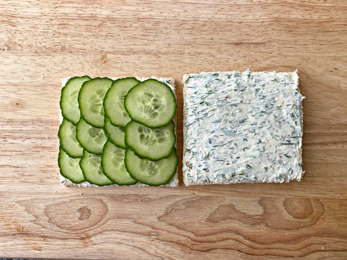 Two pieces of white bread open face, on one side are 12 slices of neatly placed cucumbers. On the other side is a piece of bread coated with the cream cheese filling.
