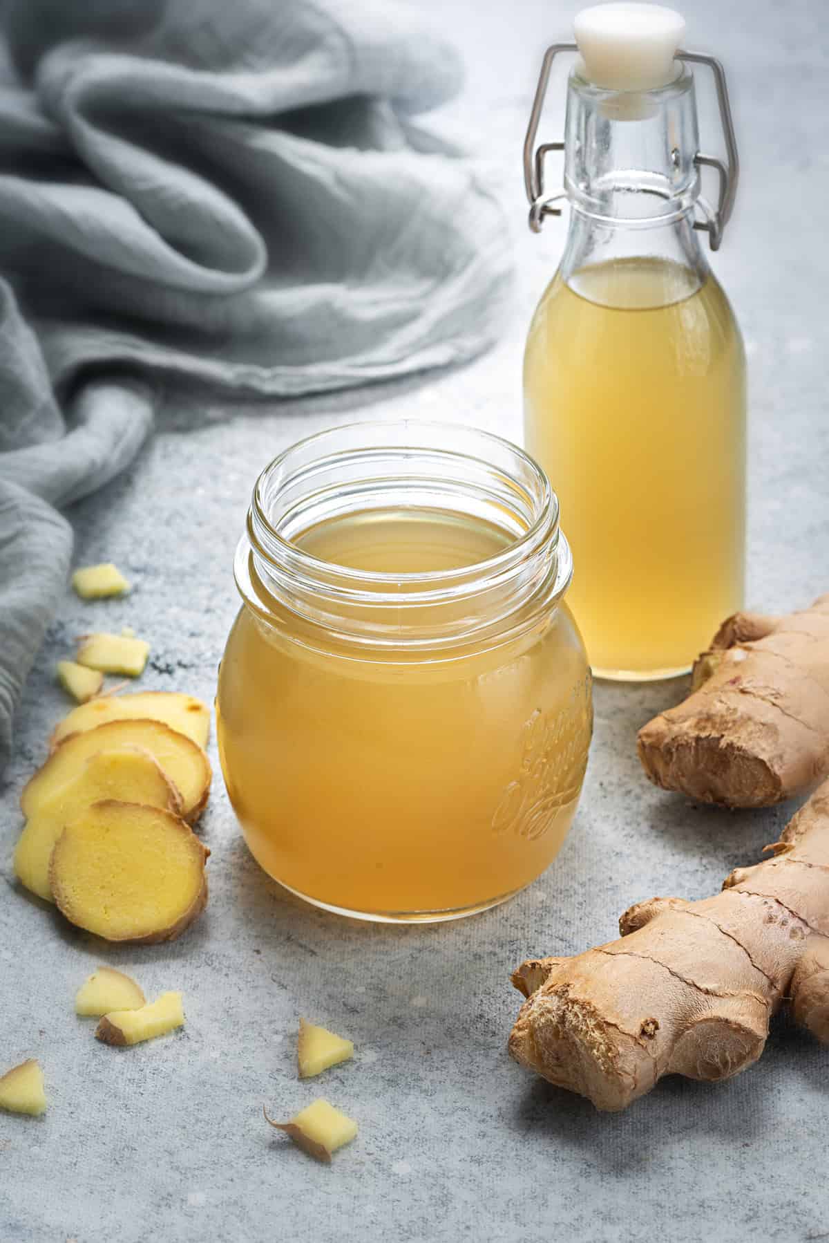 Two glass containers of simple syrup with ginger surrounded by ginger root.