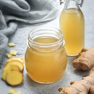 Two glass containers of simple ginger syrup with ginger surrounded by ginger root.