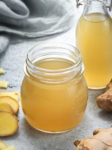 Two glass containers of simple ginger syrup with ginger surrounded by ginger root.
