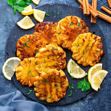 Grilled pineapple slices on a slate grey surface surrounded by lemon slices and cinnamon sticks.