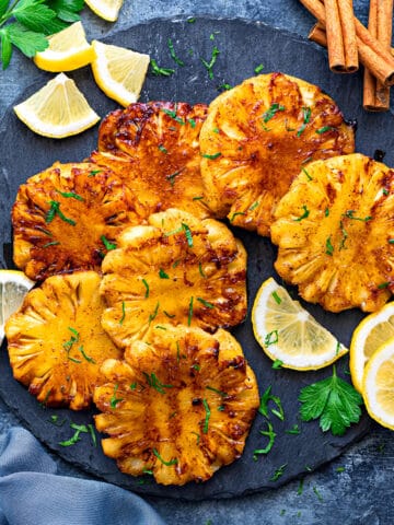 Grilled pineapple slices on a slate grey surface surrounded by lemon slices and cinnamon sticks.