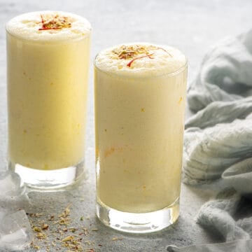 Two tall glasses of sweet lassi topped with nuts and saffron strands.