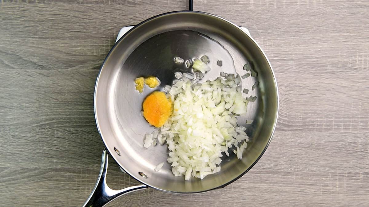 Top down view deep metal skillet with minced garlic and diced onion in oil.