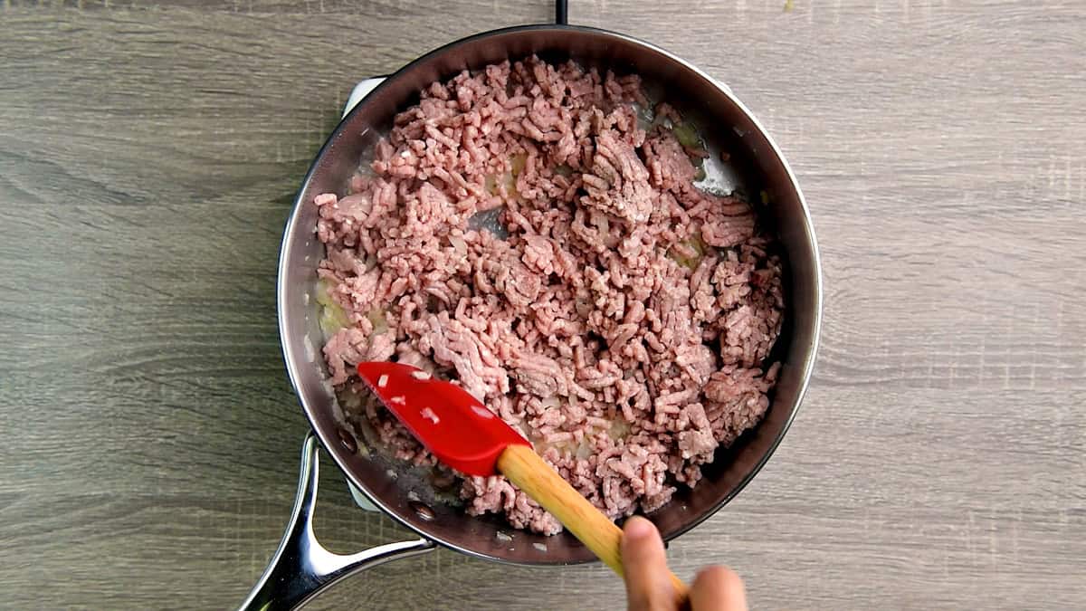 Top down view deep metal skillet with ground beef being stirred by a red spatula.