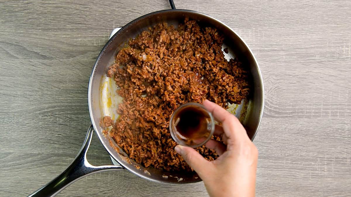 Hand adding Worcestershire sauce from a small glass bowl into a pan of cooked ground beef.