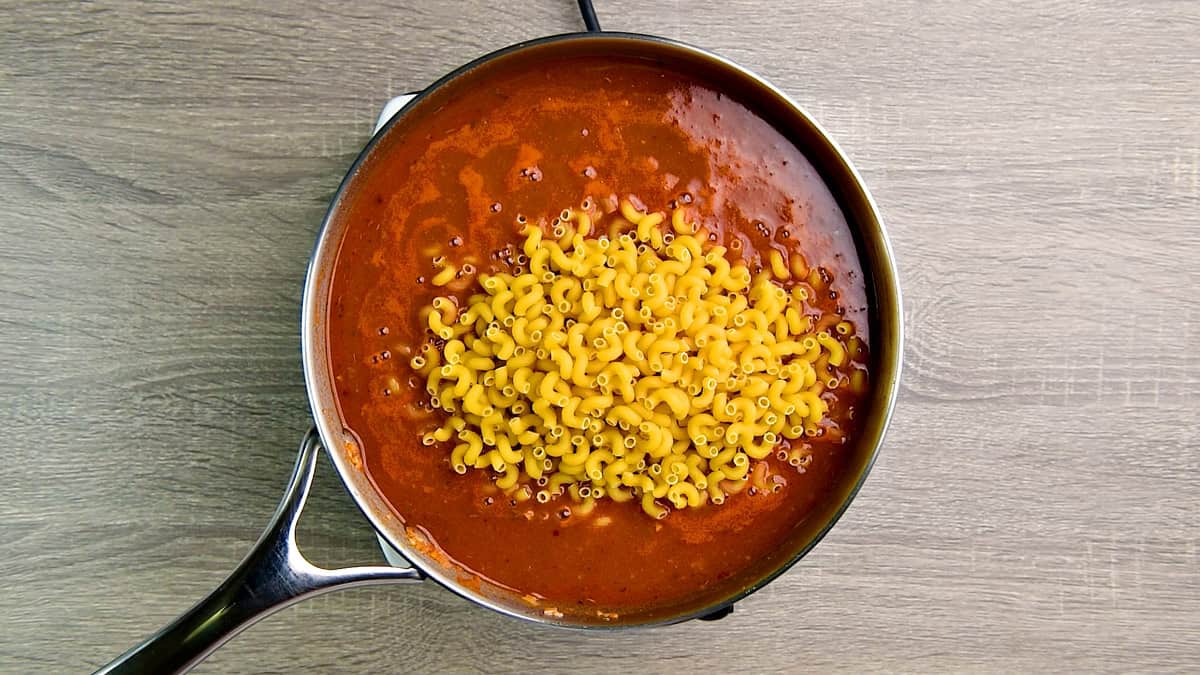 Adding elbow macaroni noodles to a skillet filled with ground beef, tomato sauce and chicken broth.