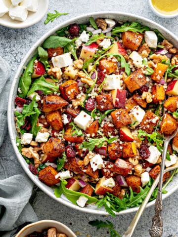 A bowl of arugula butternut squash and quinoa salad, with cubes of feta and apple.