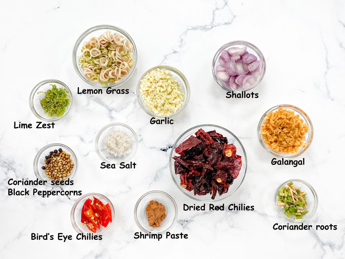 Top-down view of Thai red curry paste ingredients in individual glass bowls laid on a white table.