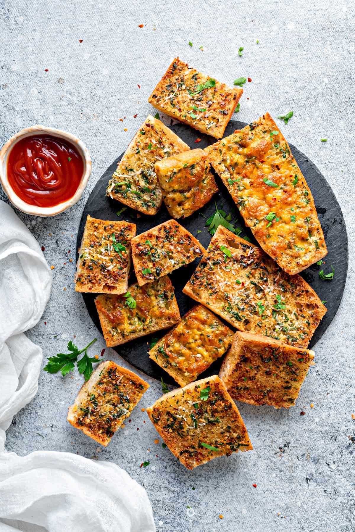 Top-down view of air fried garlic bread on a black slate topped with parsley and shredded parmesan, with a bowl of ketchup on side.