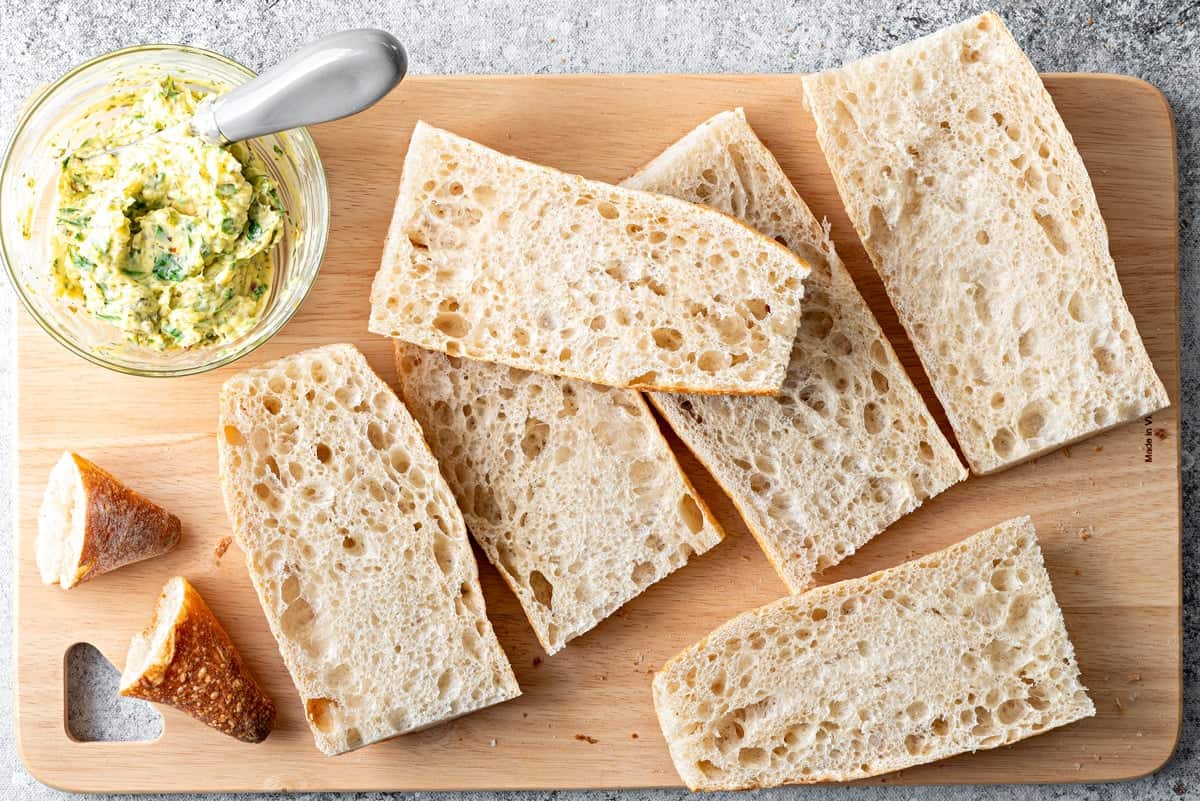 Top-down view of garlic butter mixture and baguette that has been cut into 3rds and halved on a cutting board.