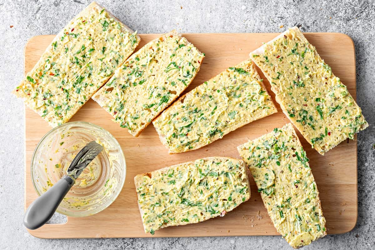 Top-down view of garlic butter mixture that has been spread on a baguette that has been cut into 3rds and halved on a cutting board.