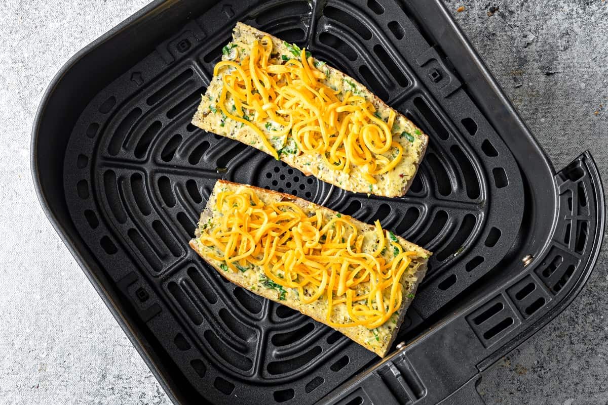 Top-down view of garlic bread topped with cheddar cheese that is in an air fryer, ready to be cooked.