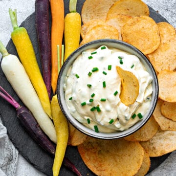 Top-down view of sour cream dip with chives in a bowl on a plate surrounded by chips and carrots for dipping and enjoying.