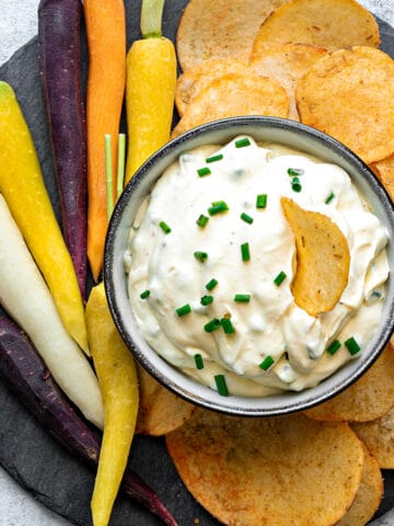 Top-down view of sour cream dip with chives in a bowl on a plate surrounded by chips and carrots for dipping and enjoying.