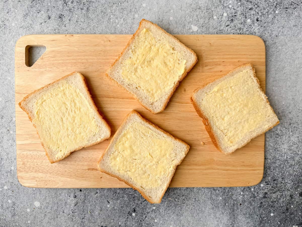 Top-down view of four whit bread slices smeared with butter on a wooden cutting board.