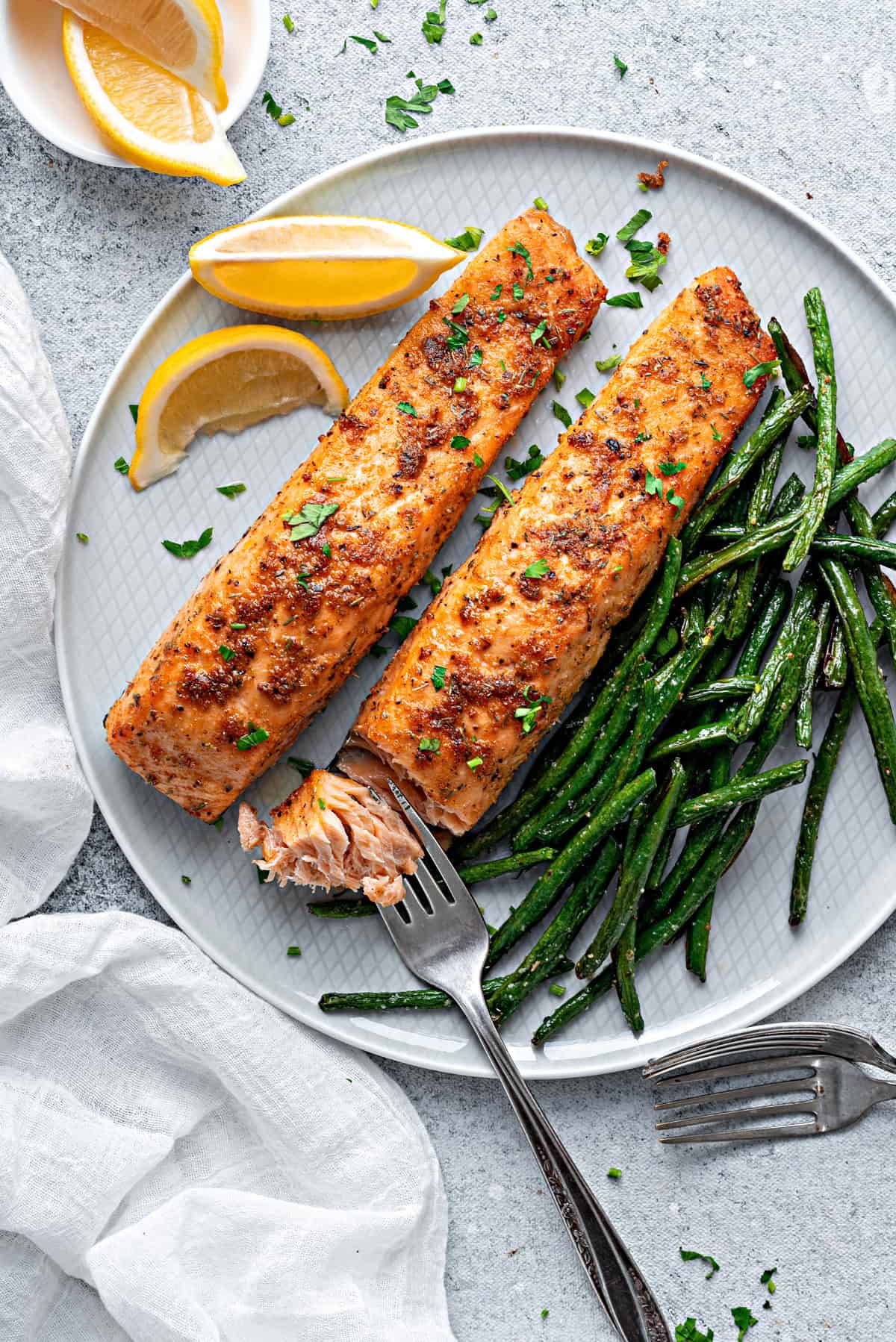 A close-up of air-fried salmon filets flaked with a fork on a plate with green beans and lemon garnish served for dinner.