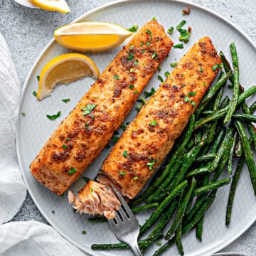 A close-up of air-fried salmon filets flaked with a fork on a plate with green beans and lemon garnish served for dinner.
