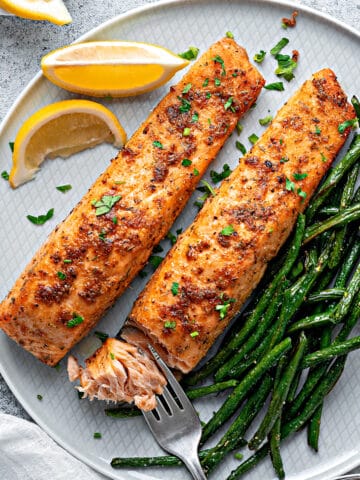 A close-up of air-fried salmon filets flaked with a fork on a plate with green beans and lemon garnish served for dinner.