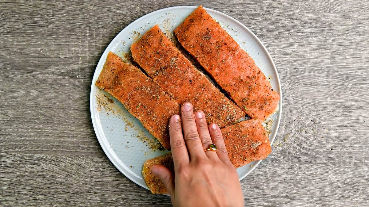 Top down view of a hand patting and spreading the spices evenly over the salmon fillets.