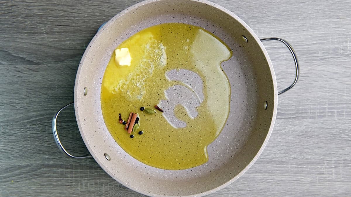A heavy-bottomed skillet that has green cardamom, cinnamon stick, cloves, and black peppercorns cooking in a mix of butter and olive oil.