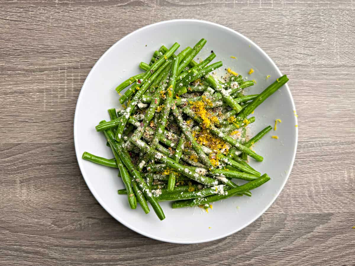 Olive oil coated green beans in a bowl that are topped with Italian seasoning, garlic, lemon zest, salt and pepper.
