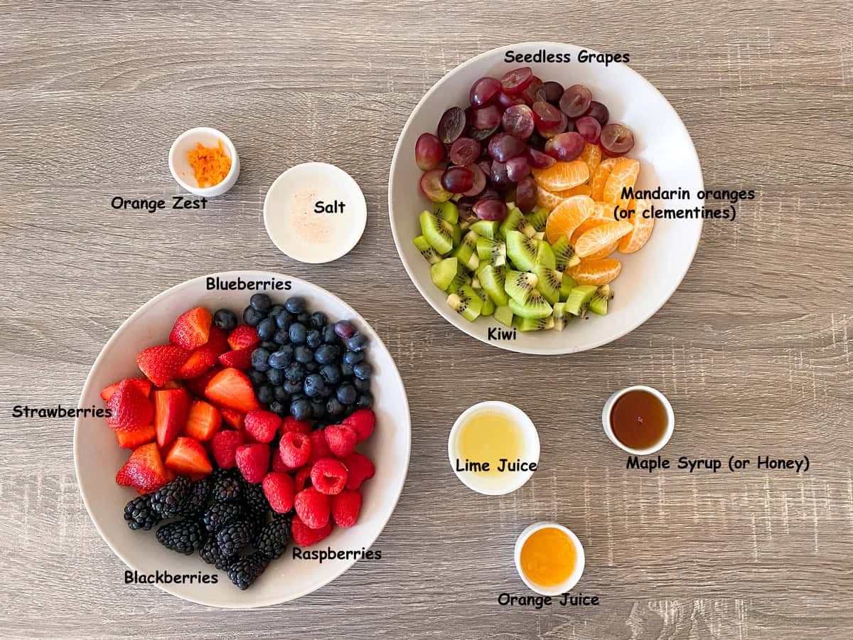 Recipe ingredients in glass bowls for berry fruit salad that include strawberries, blackberries, kiwi, grapes, lime juice, salt, orange juice and more.