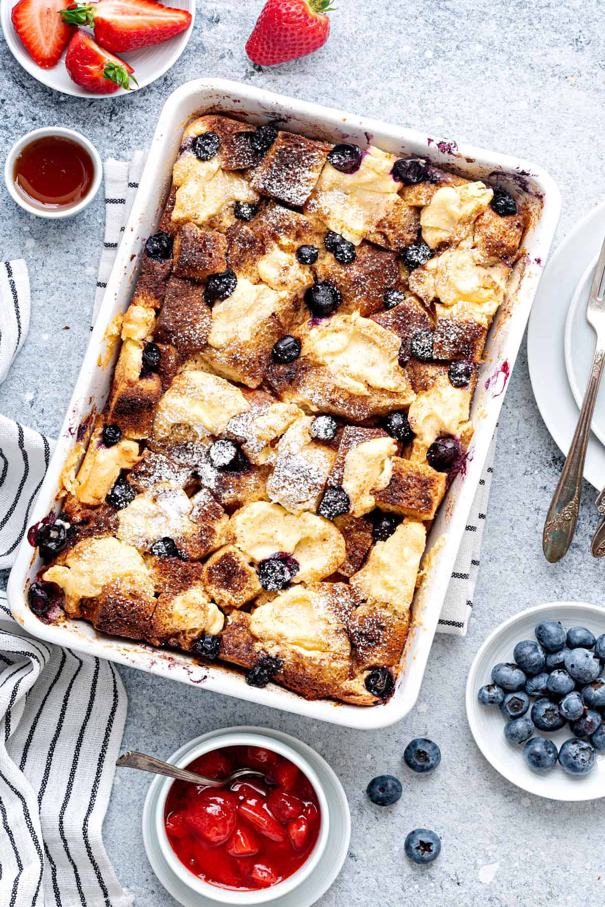 Baked Breakfast brioche french toast casserole in a 9x13 baking dish sprinkled with confectioner sugar. Blueberries and maple syrup on side.