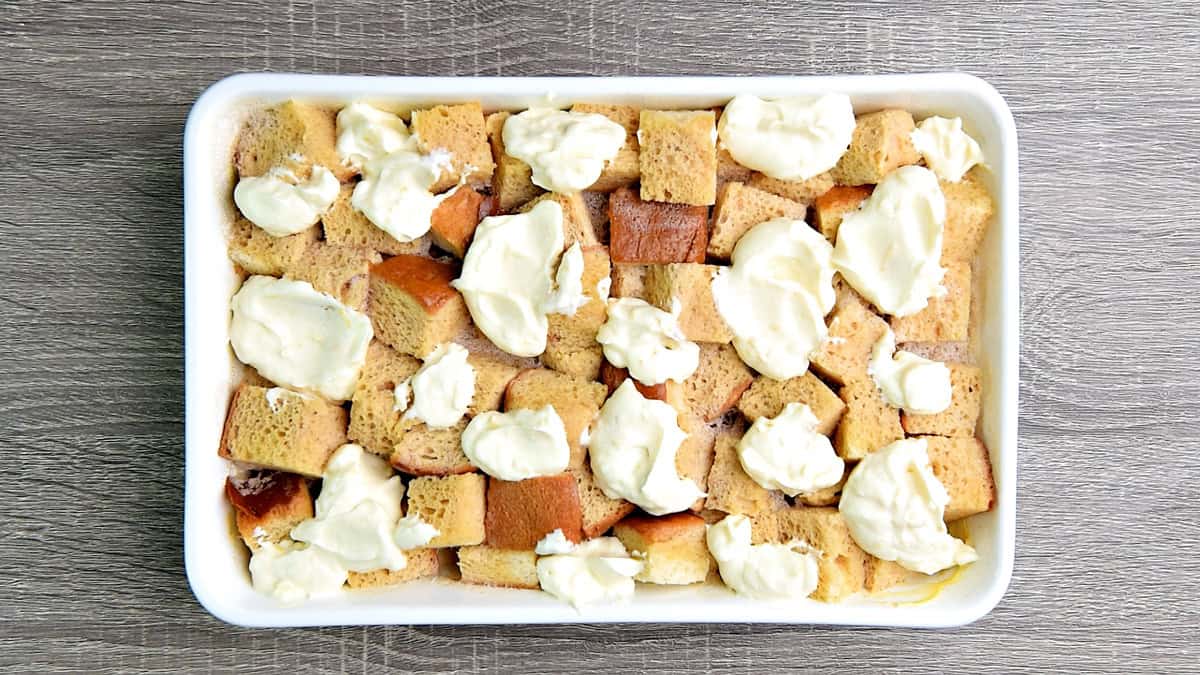 The refrigerated baking dish with cubed brioche and custard uncovered and dolloped with prepared creamy cheesecake mixture.